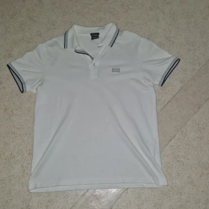 Vit pikétröja från Hugo Boss - Vit pikétröja från Hugo Boss med klassisk krage och knäppning framtill. Tröjan har mörka detaljer på krage och ärmslut samt broderad logga på bröstet. Tillverkad i bomull och har en regular fit som sitter snyggt och bekvämt.