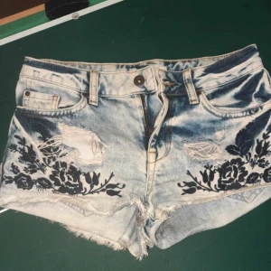 Blå jeansshorts med broderi Greencoast - Snygga ljusblå jeansshorts från Greencoast med slitna detaljer och svarta blommiga broderier framtill. Shortsen har fransig kant, klassisk femficksmodell och knappgylf. Perfekta för sommaren och ger en cool vibe till din outfit.