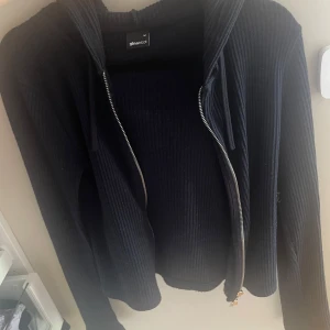 Svart ribbad cardigan från Gina Tricot - Svart ribbad hoodie från Gina Tricot med dragkedja framtill och huva. Tröjan har långa ärmar och är tillverkad i ett mjukt stickat material som känns både bekvämt och stilrent. Perfekt för lager-på-lager och chill dagar.