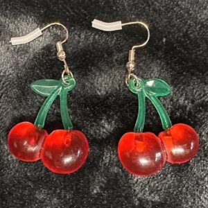 Körsbärsörhängen i röd plast – lekfull retrostil, allergisäkra krokar, helt nya - Färgglada örhängen med körsbär i transparent röd plast och gröna blad – perfekt för en retroinspirerad look eller Y2K-stil. Ca 4 cm långa, med nickelsäkra krokar. Helt nya och oanvända! Lätta att bära och riktigt blickfångande. Ingen prutning – men jag erbjuder bundlerabatt. Rensar både smyckesskrin och garderob, så passa på att kika på mina andra annonser också!  ⸻  