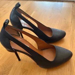 Svarta pumps med spetsig tå - Snygga svarta pumps från other stories i skinn med spetsig tå och hög klack. Skorna har en öppen design på sidorna och en elegant rem med ögla bak. Tillverkade i skinn med en stilren look som passar perfekt till trendiga outfits.