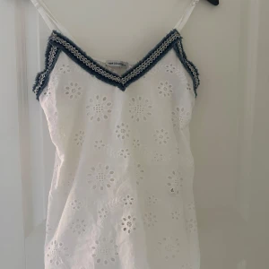 Vit broderad linnetopp från New Collection - Superfin vit linnetopp med broderade blommor och spetsdetaljer. Toppen har smala axelband och v-ringning med blå och vit spetskant. Perfekt för en somrig look och lätt att matcha med jeans eller kjol.