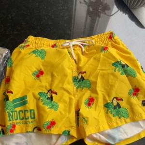 Gula badshorts med tropiskt tryck NOCCO - Färgglada gula badshorts från NOCCO med tropiskt tryck av tukaner, gröna blad och röda blommor. Shortsen har elastisk midja med vit snörning och är perfekta för strand eller pool. Snygg logga på vänster ben och snabbtorkande material. Det är dom förtsa nocco shortsen som tilvirkades så dom går inte att få tag på. dom är i storlek L men är lite små för sin storlek