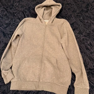Grå hoodie från ONLY & SONS med dragkedja - Säljer en stilren grå hoodie från ONLY & SONS i storlek M. Tröjan har hel dragkedja, huva med snörhål och två fickor framtill. Perfekt för chill dagar eller när du vill ha en enkel och clean look.