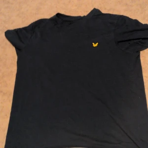 Svart t-shirt från Lyle & Scott - Svart t-shirt från Lyle & Scott med klassisk gul fågel-logga på bröstet. Enkel och stilren design, rund hals och korta ärmar. Tillverkad i en mjuk blandning av bomull och polyester som känns skön mot huden.