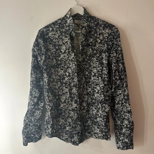 Eton skjorta  - Svart skjorta med ett blommigt mönster i vitt och grått. Skjortan har långa ärmar och en klassisk krage. Slim fit modell. Storlek 38/S