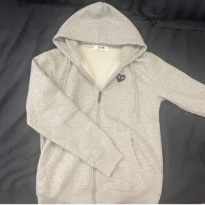 Grå hoodie Comme des Garçons Play  - Grå hoodie från Comme des Garçons Play med dragkedja och huva. Ikoniskt svart hjärta med ögon på bröstet och stort tryck på ryggen. Mjuk bomullskänsla och klassisk passform, perfekt för en chill och stilren look.