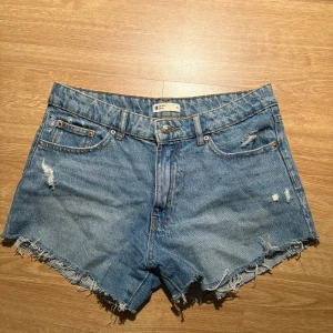 Blå jeansshorts med slitningar - Snygga blå jeansshorts från Gina Tricot med råa, fransiga kanter och slitna detaljer framtill. Klassisk femficksmodell med knapp och dragkedja. Perfekta för varma dagar och en avslappnad stil.