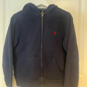 Mörkblå hoodie från Polo Ralph Lauren - Snygg mörkblå hoodie från Polo Ralph Lauren med dragkedja och klassisk röd logga på bröstet. Tröjan har huva, känguruficka och är tillverkad i mjuk bomull. Perfekt för en chill och stilren look.