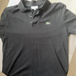 Svart pikétröja Lacoste slim fit XL - Klassisk svart pikétröja från Lacoste i slim fit-modell. Tröjan har korta ärmar, krage med två knappar och den ikoniska gröna krokodilloggan broderad på bröstet. Tillverkad i bomull för en skön och luftig känsla. Skick 9/10, passar Large.
