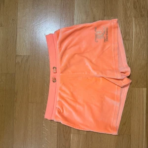 Juicy Couture peachfärgade shorts - Supermjuka peachfärgade shorts från Juicy Couture i velour med bred resår i midjan och snörning. Ikonisk logga med strassdetaljer på ena benet. Perfekta för chill eller sommarhäng.