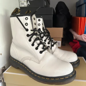 Vita Dr. Martens boots med svarta snören - Säljer ett par klassiska vita Dr. Martens boots med svarta snören och gul söm runt sulan. Skorna har rund tå, platt sula och är tillverkade i skinn. Perfekta för dig som vill sticka ut med en ikonisk stil.