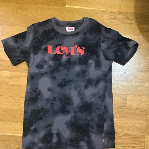 Svart tie-dye Levi's t-shirt - Snygg svart och grå tie-dye t-shirt från Levi's med röd logga framtill. Klassisk passform med korta ärmar och rund halsringning. Perfekt för dig som gillar streetstyle och vill sticka ut lite extra.