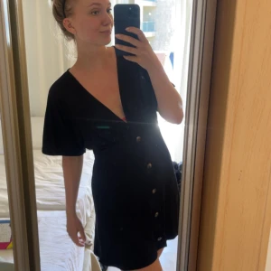 Svart kortklänning med knappar - Svart kortklänning med djup v-ringning och knappar framtill. Klänningen har korta ärmar och en avslappnad passform som ger en snygg och enkel look. Perfekt som strandklänning i mjukt material.