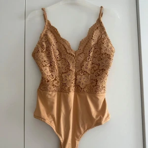 Beige spets body med smala axelband - Supersnygg beige body  med smala axelband och v-ringning. Överdelen är täckt av blommig spets med fina detaljer och nederdelen är i mjukt tyg. Perfekt för att styla med jeans eller kjol för en trendig look