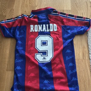 Barcelona Ronaldo 9 Kappa fotbollströja - Retro Barcelona fotbollströja med Ronaldo 9 på ryggen. Klassiska blå och röda vertikala ränder, tryckt Barca- och Kappa-logga över hela tröjan samt klubbmärke på bröstet. Kortärmad modell i lätt och ventilerande polyester, perfekt för dig som älskar vintage och fotbollshistoria. Tröjan är i storlek medium, kan diskutera pris!