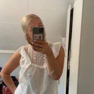 Vit blus med spets och volang - Supersöt vit blus med broderad spets upptill och volangdetaljer längs axlarna. Ärmlös modell med romantisk känsla och luftig passform. Perfekt för dig som gillar en feminin och trendig stil.