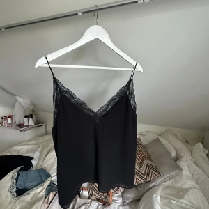 Svart linnetopp med spetskant - Svart linnetopp med tunna axelband och v-ringning dekorerad med spets. Snygg och enkel modell som passar perfekt till jeans eller kjol. Toppens material känns lätt och luftigt, och spetsdetaljen ger en extra touch.