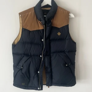 Svart och brun dunväst från Morris - Snygg dunväst från Morris i svart med bruna detaljer i manchester på axlarna. Västen har hög krage, dragkedja och tryckknappar framtill samt två fickor med lock. Perfekt för lager-på-lager och en cool look.