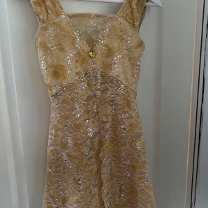 Kondtåkningsklänning   - Superfin tävlingsklänning i champagnefärg med blommig spets, paljetter och v-ringning både fram och bak. Klänningen har breda axelband och är figursydd upptill med lätt utställd kjol. Den är skärddarsydd och har flera swarovskidiamanter
