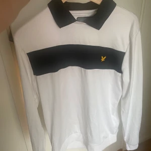Vit och svart pikétröja från Lyle & Scott - Snygg långärmad pikétröja från Lyle & Scott i vitt med svart krage och svart panel över bröstet. Klassisk gul logga broderad på bröstet. Perfekt för dig som gillar stilren och sportig look.