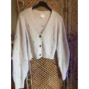 Beige kofta med broderade blommor H&M - Superfin beige kofta från H&M med broderade små blommor i pastellrosa och grönt. Koftan är croppad, har v-ringning, stora knappar och långa puffiga ärmar. Materialet är en mjuk mix av polyester, akryl, ull och elastan. Perfekt för dig som gillar en söt och trendig look.