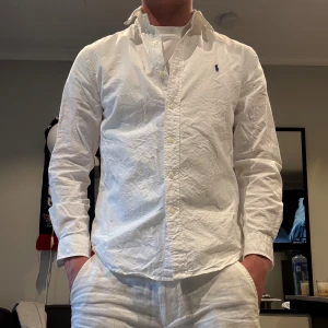 Vit skjorta från Ralph Lauren - Klassisk vit skjorta från Ralph Lauren med button-down krage och broderad logga på bröstet. Skjortan är långärmad, har normal passform och är tillverkad i bomull. Perfekt för en clean och stilren look.