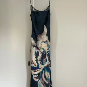 Långklänning med blommigt mönster - Snygg långklänning med tunna axelband och stor blommig print i blått, beige och grått. Klänningen har öppen rygg med snörning och är gjord i ett glansigt, satinliknande material. Perfekt för dig som vill sticka ut med en unik och elegant look.