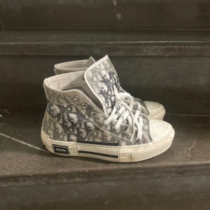 Dior high-top sneakers med monogram - Snygga high-top sneakers från Dior med genomskinlig ovandel och klassiskt grått Dior-monogram. Skorna har vita snören, rund tå och platt sula med Dior-logga på sidan och unika bi-detaljer under sulan. Perfekt för dig som vill sticka ut med lyxig streetstyle. Köpte på Vinted, äkta, har tagit ut sulan i. 