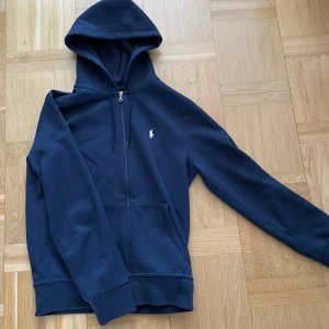 Marinblå hoodie från Polo Ralph Lauren - Snygg marinblå hoodie från Polo Ralph Lauren med dragkedja och klassisk broderad logga på bröstet. Tröjan har huva med snörning, känguruficka och är tillverkad i mjuk bomullsmix. Perfekt för en chill och stilren look.