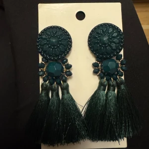 Mörkgröna örhängen med tofsar - Snygga mörkgröna örhängen med rund dekorerad topp, glittrande stenar och hängande tofsar i textil. Helt oanvända.