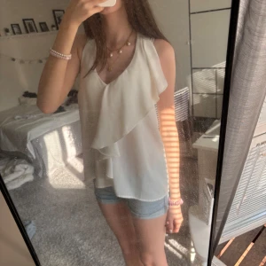 Vit linnetopp med volang - Säljer en superfin vit linnetopp volangdetaljer framtill. Toppen är ärmlös och har en snygg v-ringning, perfekt för varma dagar. Lätt och luftig i materialet, passar till både jeans och kjol.