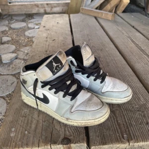 Nike Air Jordan 1 Mid vit/svart EUR 38 - Säljer ett par Nike Air Jordan 1 Mid i vitt och svart med klassisk svart swoosh och svarta skosnören. Skorna har hög siluett, perforerad tå och ikoniska Air Jordan-loggan på sidan. Ovandelen är i skinn och de har en platt sula. Perfekta för dig som gillar streetstyle.