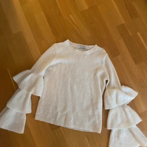 Vit stickad tröja med volangärmar NA-KD - Superfin vit stickad tröja från NA-KD med unika volangärmar i tre lager. Tröjan har rund halsringning och ribbade muddar. Perfekt för dig som vill sticka ut med en trendig och mysig look.