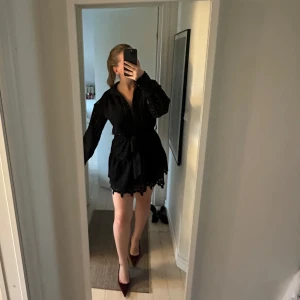 Svart kort klänning med knytband - Supersnygg svart kort klänning med långa ärmar och knytband i midjan. Klänningen har broderade detaljer och volangkant nertill som ger en extra touch. Perfekt för dig som vill ha en stilren men ändå unik look.