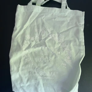 Vit ljus grön tygkasse från COCO-MAT - Säljer en vit ljus grön shoppingväska i bomull från COCO-MAT med trycket 'natural all the way' och blommiga detaljer. Väskan har två långa handtag och är perfekt för att bära böcker, mat eller annat du behöver under dagen.