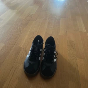 Svarta Adidas sneakers med vita ränder - Helt nya och oanvända, Snygga svarta Adidas sneakers med klassiska vita ränder på sidorna och svart sula. Skorna har snörning och är tillverkade i en mix av syntet och skinn. Perfekta för dig som gillar en clean och sportig look.