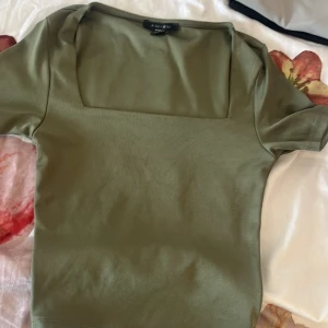 Olivgrön topp med fyrkantig ringning - Snygg olivgrön topp från Amisu med korta ärmar och trendig fyrkantig ringning. Perfekt att matcha med jeans eller kjol för en clean och stilren look. Mjuk och skön bomullskänsla som sitter bra på kroppen.