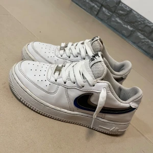 Nike Air Force 1 vita sneakers - Nike Air Force 1 sneakers i vitt skinn med svart och blå swoosh på sidan. Klassisk låg modell med perforerad tå, vita snören och grå meshfoder. Perfekta för dig som gillar streetstyle och vill ha en ikonisk sneaker i garderoben.