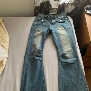 Blå bootcut jeans med råa kanter - Säljer ett par blå bootcut jeans med coola råa kanter nertill och stora fickor framtill med vita knappar. Jeansen har en ljus tvätt och markerade sömmar, samt dragkedja i gylfen. Perfekta för dig som gillar en retro vibe och vill sticka ut.