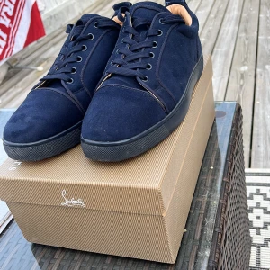 Mörkblå sneakers från Louboutin - Snygga mörkblå sneakers från Christian Louboutin i mocka med klassisk röd sula och svarta detaljer. Skorna har rund tå, snörning och en stilren design som sticker ut med den ikoniska röda undersidan. Perfekta för dig som vill ha något exklusivt och trendigt.