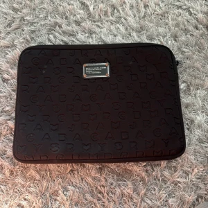 Svart datorväska Marc Jacobs - Säljer en svart datorväska från Marc Jacobs med präglade bokstäver över hela ytan och en metallbricka framtill. Dragkedjan är har ”hoppat av” men går säkert att fixa. 