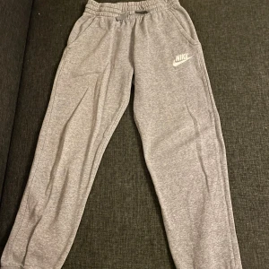 Grå mjukisbyxor från Nike L - Snygga grå mjukisbyxor i bomull från Nike med broderad logga på benet. Byxorna har resår och snörning i midjan samt sidofickor. Perfekta för chill eller träning, tillverkade i mjukt material för maximal komfort.