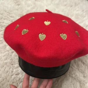 Röd basker med guldhjärtan - Superfin röd basker i ull med broderade guldhjärtan över hela ovansidan. Klassisk fransk form med liten topp och snygg detalj med hjärtan som ger en lekfull touch. Perfekt accessoar för att sticka ut och addera färg till din look.