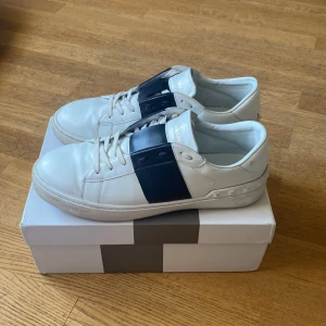 Valentino Garavani Open sneakers vit/blå - Snygga Valentino Garavani Open sneakers i vitt skinn med bred marinblå rem över vristen och coola nitar på hälen. Klassisk rund tå, vita snören och platt sula. Perfekta för dig som vill ha en stilren men ändå unik look.