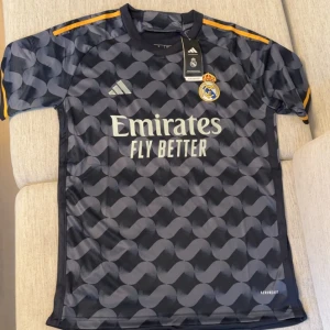 Real Madrid bortatröja Adidas svart - Snygg Real Madrid bortatröja från Adidas i svart med grått grafiskt mönster. Kortärmad modell med gula detaljer på axlarna och klubbmärke på bröstet. Tillverkad i lätt och ventilerande polyester med Aeroready-teknologi. Perfekt för dig som vill sticka ut på planen.