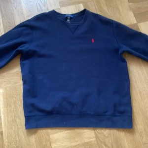 Mörkblå sweatshirt från Polo Ralph Lauren - Klassisk mörkblå sweatshirt från Polo Ralph Lauren i storlek XL. Tröjan har rund halsringning, långa ärmar och ikoniska röda broderade loggan på bröstet. Perfekt för en chill och stilren look.