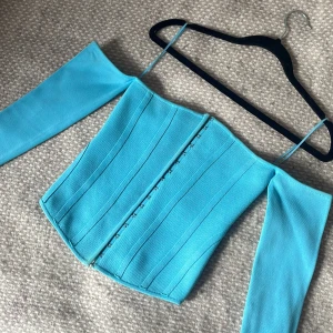 Turkos korsett-topp med långa ärmar - Säljer en snygg turkos korsett-topp med offshoulder-långa ärmar. Toppen har markerade sömmar och stängs framtill med små hyskor. Materialet är stretchigt och känns som stickad syntet. (De tunna banden är inte axelband).
