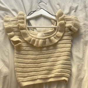 Beige stickad topp med volang - Supersöt beige stickad topp med korta puffärmar och volangdetaljer vid halsringningen och axlarna. Ribbad struktur och croppad passform gör den perfekt till både jeans och kjol. Perfekt för vår och sommar!