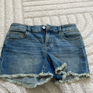 Blå jeansshorts med fransig kant - Snygga blå jeansshorts med fransig kant nertill och klassisk femficksdesign. Perfekta för varma dagar och ger en avslappnad vibe. Shortsen har normal passform och stängs med knapp och dragkedja framtill.